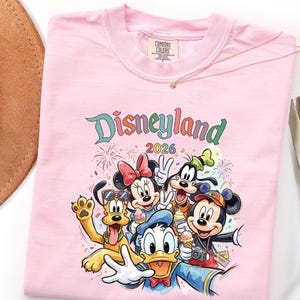 Może przedstawiać: Jasnor&oacute;żowy t-shirt z nadrukiem graficznym przedstawiającym postacie Disneya, w tym Myszkę Miki, Myszkę Minnie, Kaczora Donalda, Goofy'ego i Pluto. Nad postaciami trzymającymi rożki z lodami znajduje się napis "Disneyland 2026".