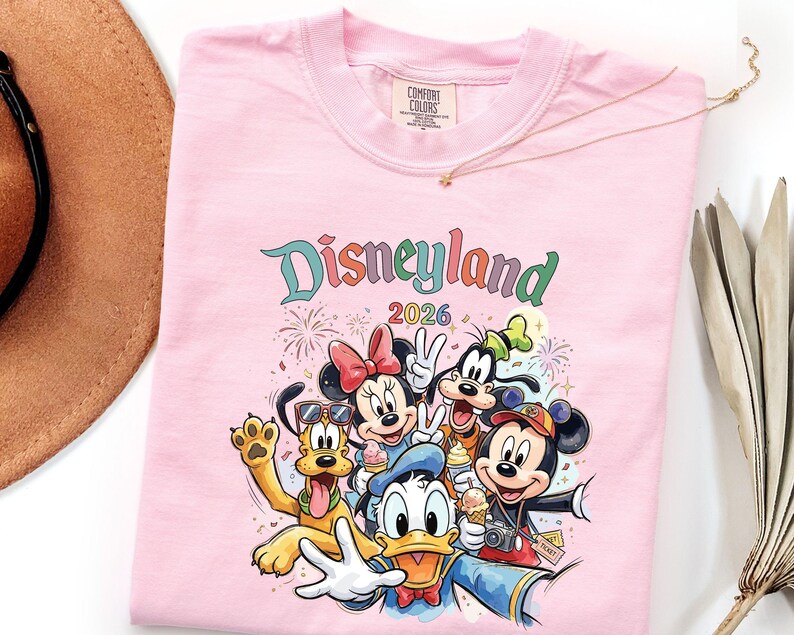 Może przedstawiać: Jasnor&oacute;żowy t-shirt z nadrukiem graficznym przedstawiającym postacie Disneya, w tym Myszkę Miki, Myszkę Minnie, Kaczora Donalda, Goofy'ego i Pluto. Nad postaciami trzymającymi rożki z lodami znajduje się napis "Disneyland 2026".