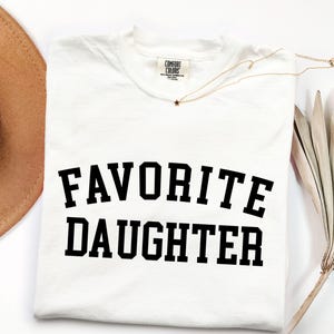 Peut inclure: T-shirt blanc avec l'inscription "FAVORITE DAUGHTER" en lettres noires grasses. Le t-shirt a un col rond et une &eacute;tiquette Comfort Colors. Un collier dor&eacute; avec un pendentif en forme d'&eacute;toile et un chapeau de cowboy beige sont &eacute;galement visibles.