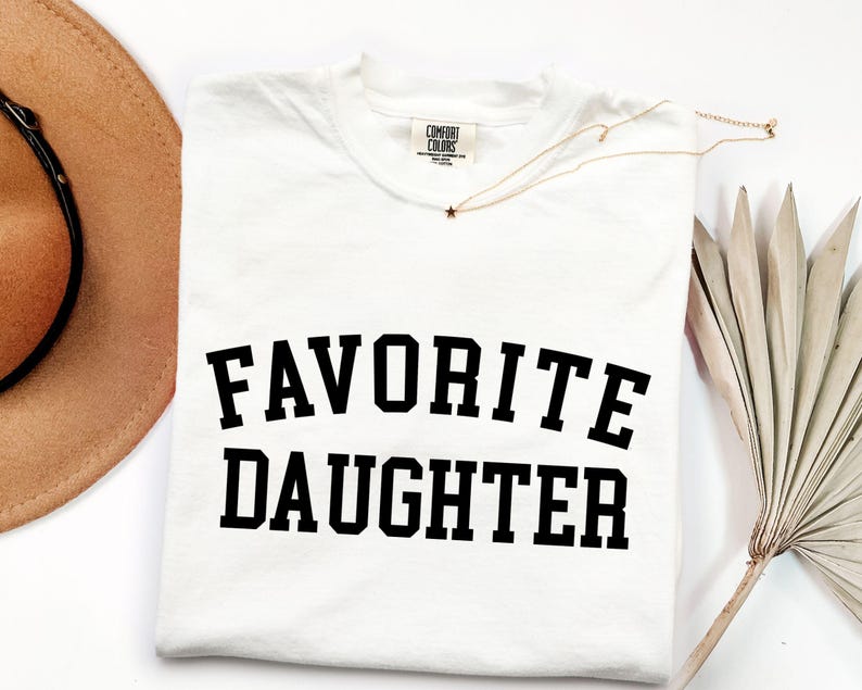 Peut inclure: T-shirt blanc avec l'inscription "FAVORITE DAUGHTER" en lettres noires grasses. Le t-shirt a un col rond et une &eacute;tiquette Comfort Colors. Un collier dor&eacute; avec un pendentif en forme d'&eacute;toile et un chapeau de cowboy beige sont &eacute;galement visibles.