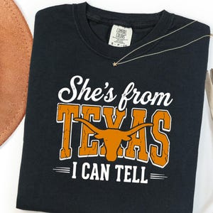 Pu&ograve; includere: Una t-shirt nera a maniche corte con una stampa grafica bianca e arancione. Il testo recita "She's from TEXAS I CAN TELL" con una silhouette di un toro longhorn arancione integrata nella parola Texas. La maglietta ha un'etichetta Comfort Colors.