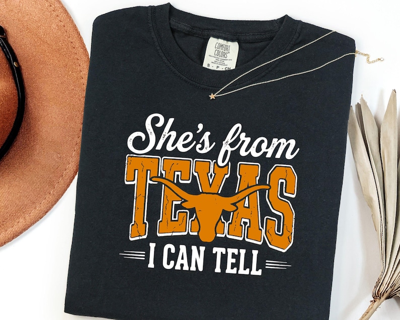 Pu&ograve; includere: Una t-shirt nera a maniche corte con una stampa grafica bianca e arancione. Il testo recita "She's from TEXAS I CAN TELL" con una silhouette di un toro longhorn arancione integrata nella parola Texas. La maglietta ha un'etichetta Comfort Colors.