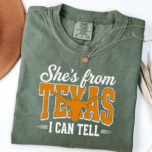 Pu&ograve; includere: Una t-shirt a maniche corte verde oliva con testo bianco e arancione che recita She's from TEXAS I CAN TELL. Il design include la sagoma arancione di un bue longhorn. La maglietta presenta un girocollo e un'etichetta con taglia S.