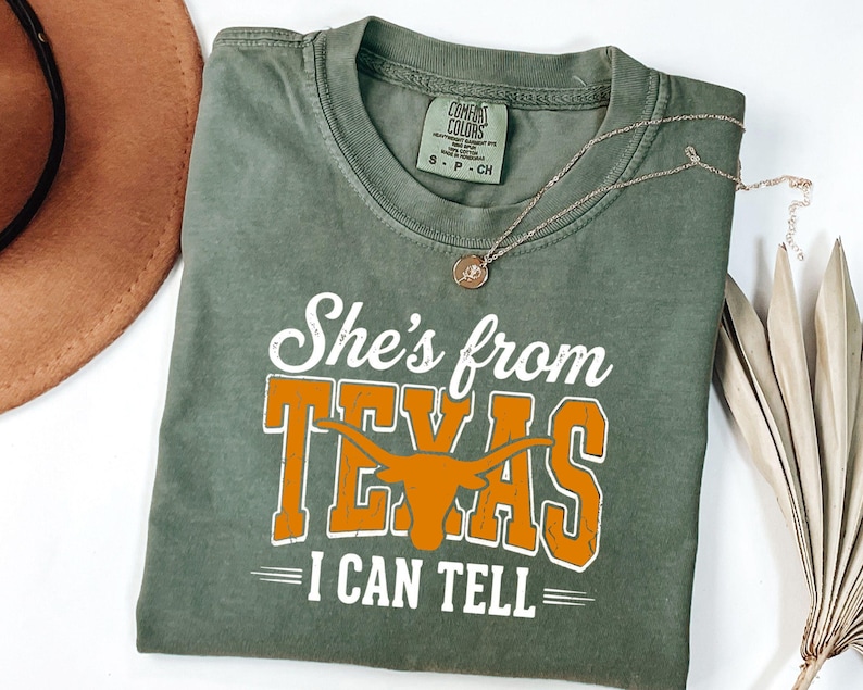 Pu&ograve; includere: Una t-shirt a maniche corte verde oliva con testo bianco e arancione che recita She's from TEXAS I CAN TELL. Il design include la sagoma arancione di un bue longhorn. La maglietta presenta un girocollo e un'etichetta con taglia S.