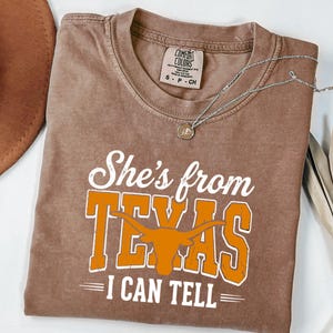 Pu&ograve; includere: Una t-shirt marrone a maniche corte con il testo bianco e arancione She's from TEXAS I CAN TELL e una grafica di una testa di toro longhorn. La maglietta presenta un girocollo e un'etichetta del marchio Comfort Colors visibile.