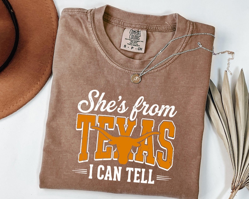 Pu&ograve; includere: Una t-shirt marrone a maniche corte con il testo bianco e arancione She's from TEXAS I CAN TELL e una grafica di una testa di toro longhorn. La maglietta presenta un girocollo e un'etichetta del marchio Comfort Colors visibile.