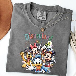 Może przedstawiać: Ciemnoszary t-shirt z okrągłym dekoltem i kolorową grafiką przedstawiającą postacie Disneya, w tym Myszkę Miki, Myszkę Minnie, Kaczora Donalda, Goofy'ego i Pluto. Nad postaciami widnieje napis Disneyland 2026. Koszulka posiada metkę Comfort Colors.