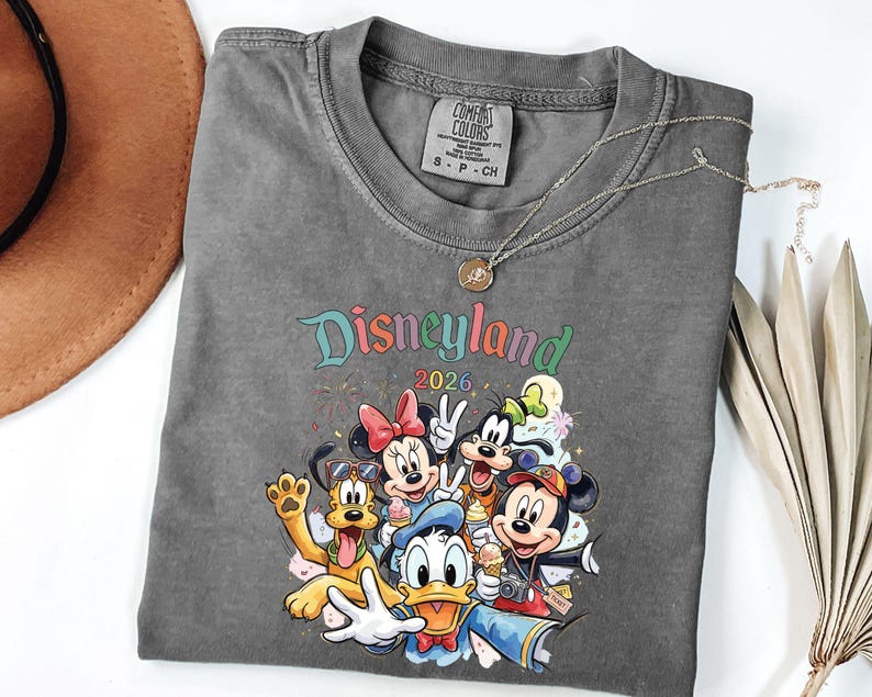 Może przedstawiać: Ciemnoszary t-shirt z okrągłym dekoltem i kolorową grafiką przedstawiającą postacie Disneya, w tym Myszkę Miki, Myszkę Minnie, Kaczora Donalda, Goofy'ego i Pluto. Nad postaciami widnieje napis Disneyland 2026. Koszulka posiada metkę Comfort Colors.