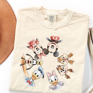 May include: Cream-coloured sweatshirt featuring a watercolour illustration of Mickey Mouse, Minnie Mouse, Donald Duck, Daisy Duck, Goofy, Chip, and Dale. The graphic is surrounded by small star accents. The shirt has a Comfort Colors tag.