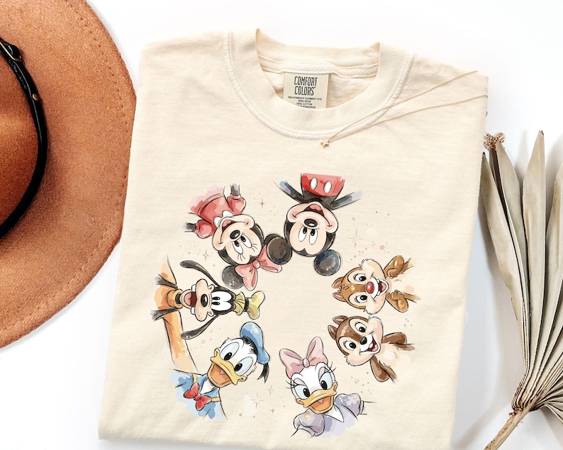 May include: Cream-coloured sweatshirt featuring a watercolour illustration of Mickey Mouse, Minnie Mouse, Donald Duck, Daisy Duck, Goofy, Chip, and Dale. The graphic is surrounded by small star accents. The shirt has a Comfort Colors tag.