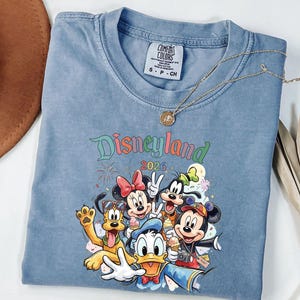 Może przedstawiać: Niebieski t-shirt z kr&oacute;tkim rękawem z grafiką przedstawiającą postacie Disneya, w tym Myszkę Miki, Myszkę Minnie, Kaczora Donalda, Goofy'ego i Pluto. Nad postaciami znajduje się napis Disneyland 2026. Koszulka posiada metkę Comfort Colors.