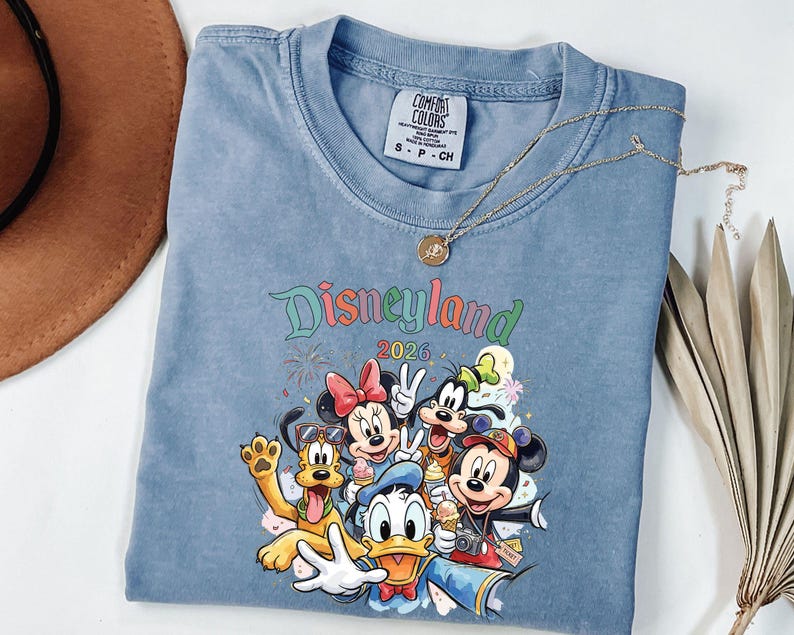 Może przedstawiać: Niebieski t-shirt z kr&oacute;tkim rękawem z grafiką przedstawiającą postacie Disneya, w tym Myszkę Miki, Myszkę Minnie, Kaczora Donalda, Goofy'ego i Pluto. Nad postaciami znajduje się napis Disneyland 2026. Koszulka posiada metkę Comfort Colors.