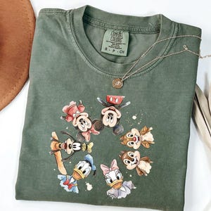May include: Green Comfort Colors t-shirt with a graphic of Mickey Mouse, Minnie Mouse, Donald Duck, Daisy Duck, Goofy, and Chip and Dale. A gold necklace with a pendant is draped across the shirt.