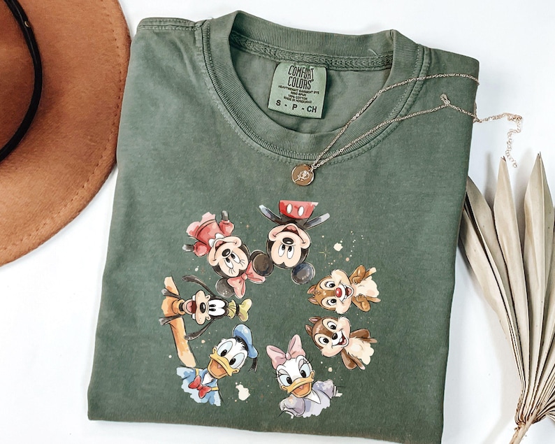 May include: Green Comfort Colors t-shirt with a graphic of Mickey Mouse, Minnie Mouse, Donald Duck, Daisy Duck, Goofy, and Chip and Dale. A gold necklace with a pendant is draped across the shirt.