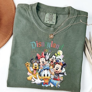 Może przedstawiać: Oliwkowozielony t-shirt z nadrukiem graficznym przedstawiającym postacie Disneya, w tym Myszkę Miki, Myszkę Minnie, Kaczora Donalda, Goofy'ego i Pluto. Nad postaciami widnieje napis Disneyland 2026. Koszulka posiada metkę marki Comfort Colors.