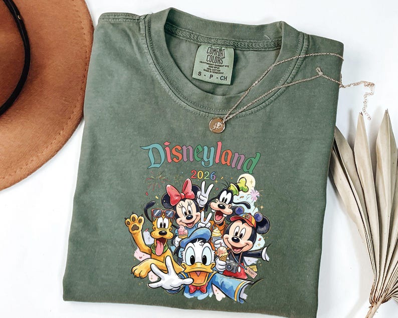 Może przedstawiać: Oliwkowozielony t-shirt z nadrukiem graficznym przedstawiającym postacie Disneya, w tym Myszkę Miki, Myszkę Minnie, Kaczora Donalda, Goofy'ego i Pluto. Nad postaciami widnieje napis Disneyland 2026. Koszulka posiada metkę marki Comfort Colors.