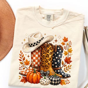 May include: A cream-colored t-shirt featuring a graphic print of a cowboy hat, three cowboy boots with checkered, leopard, and polka dot patterns, two pumpkins, and autumn leaves. The inner neck label displays the text Comfort Colors.