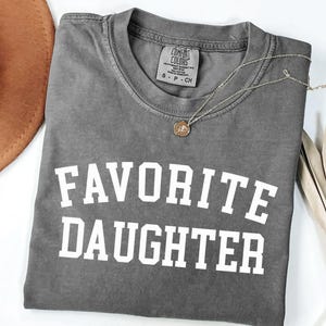 Peut inclure: Un t-shirt gris Comfort Colors avec les mots "FAVORITE DAUGHTER" en lettres blanches en gras. Un collier en or avec un pendentif repose sur le t-shirt. Un chapeau marron et des feuilles s&eacute;ch&eacute;es sont en arri&egrave;re-plan.