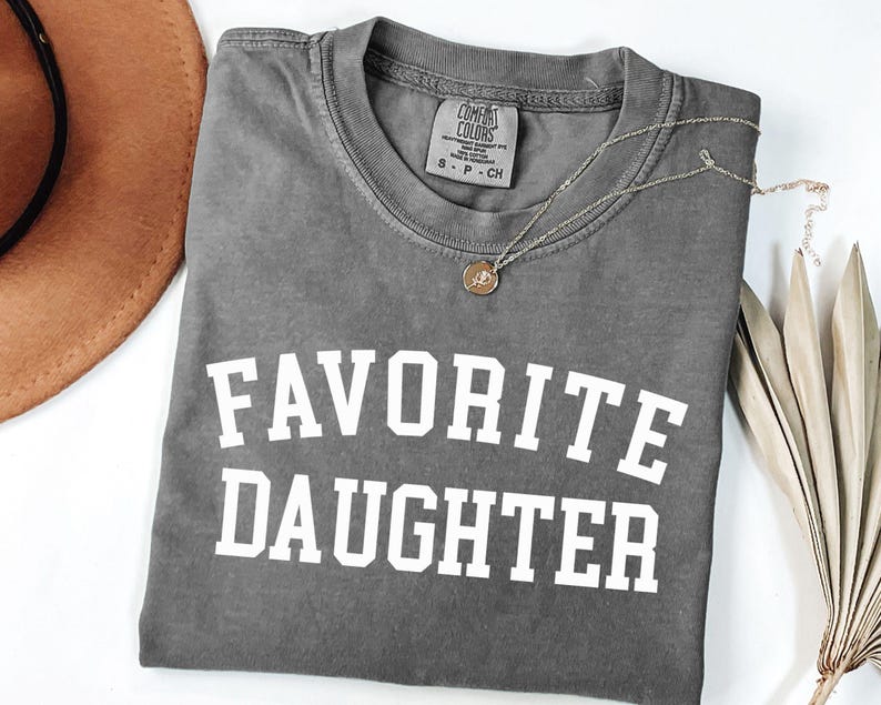 Peut inclure: Un t-shirt gris Comfort Colors avec les mots "FAVORITE DAUGHTER" en lettres blanches en gras. Un collier en or avec un pendentif repose sur le t-shirt. Un chapeau marron et des feuilles s&eacute;ch&eacute;es sont en arri&egrave;re-plan.
