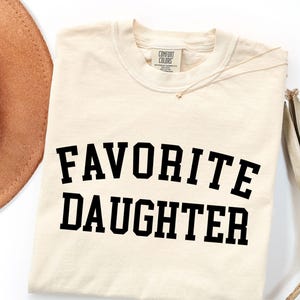 Peut inclure: T-shirt cr&egrave;me avec l'inscription "FAVORITE DAUGHTER" en lettres noires et grasses. Un collier dor&eacute; et un chapeau marron sont visibles en arri&egrave;re-plan. Le t-shirt porte une &eacute;tiquette Comfort Colors.