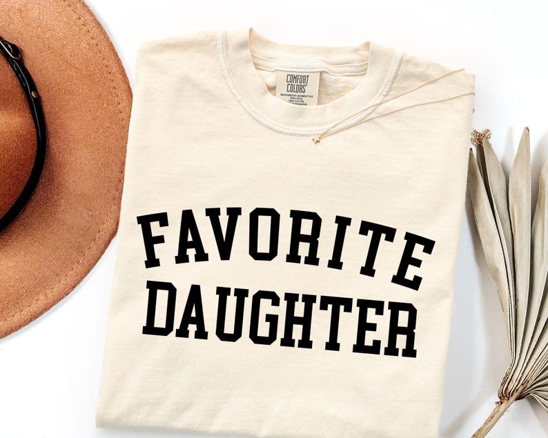 Peut inclure: T-shirt cr&egrave;me avec l'inscription "FAVORITE DAUGHTER" en lettres noires et grasses. Un collier dor&eacute; et un chapeau marron sont visibles en arri&egrave;re-plan. Le t-shirt porte une &eacute;tiquette Comfort Colors.