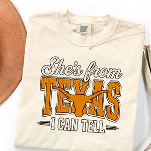 Pu&ograve; includere: Una t-shirt a maniche corte color crema con il testo "She's from TEXAS I CAN TELL" in caratteri neri e arancioni. La parola "TEXAS" include una grafica di una testa di bue longhorn arancione. La maglietta ha un'etichetta Comfort Colors.