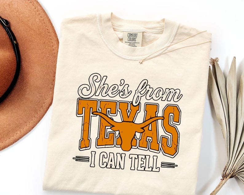 Pu&ograve; includere: Una t-shirt a maniche corte color crema con il testo "She's from TEXAS I CAN TELL" in caratteri neri e arancioni. La parola "TEXAS" include una grafica di una testa di bue longhorn arancione. La maglietta ha un'etichetta Comfort Colors.