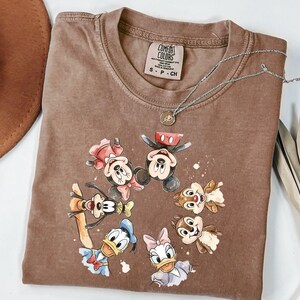 May include: Brown Comfort Colors t-shirt with a watercolour design of Mickey Mouse, Minnie Mouse, Donald Duck, Daisy Duck, Goofy, and Chip and Dale. The shirt has a crew neck and short sleeves. A silver necklace with a pendant is on the shirt.
