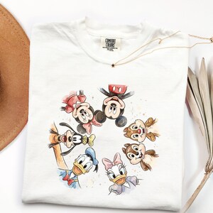 May include: White t-shirt with a watercolour illustration of Disney characters Mickey Mouse, Minnie Mouse, Donald Duck, Daisy Duck, Goofy, and Chip and Dale. The shirt has a Comfort Colors tag and a gold necklace.