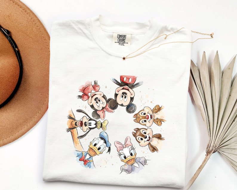 May include: White t-shirt with a watercolour illustration of Disney characters Mickey Mouse, Minnie Mouse, Donald Duck, Daisy Duck, Goofy, and Chip and Dale. The shirt has a Comfort Colors tag and a gold necklace.