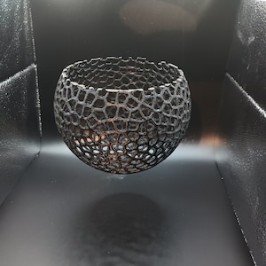 Peut inclure: Un bol sphérique noir imprimé en 3D avec un design géométrique ajouré. Le bol est suspendu en l'air, projetant une ombre sur la surface noire en dessous.