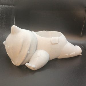 Puede incluir: Un macetero de cerámica blanca con forma de Pokémon durmiendo. El macetero tiene una pequeña abertura en la parte superior para plantar.