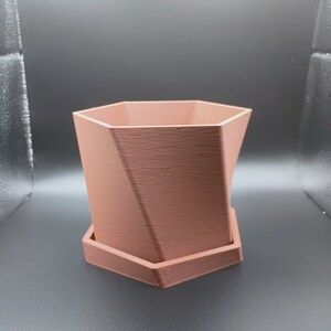 Op de afbeelding: Een terracottakleurige zeshoekige plantenpot met een bijpassende schotel. De pot heeft een geometrisch ontwerp en een gladde, matte afwerking.