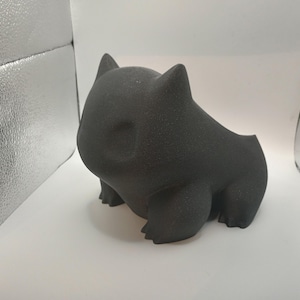 Puede incluir: Figura estilizada de color gris oscuro de una criatura con cuerpo redondeado, patas pequeñas y orejas puntiagudas. La figura tiene un acabado liso y mate, sobre un fondo blanco.
