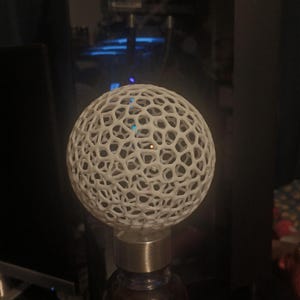 以下が含まれることがあります： 白い球状のオープン織りの装飾品がガラス瓶の上に置かれています。球体はハニカムのような模様です。ボトルは銀色のキャップで、暗い液体が入っています。背景は暗いです。