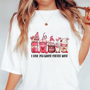 May include: White t-shirt with a graphic of five gnomes holding Valentine's Day themed drinks and the text "I love you gnome matter what".