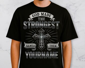 May include: Black t-shirt with the text "GOD MADE THE STRONGEST NAMED THEM YOURNAME" in a silver, ornate font. A silver cross is centered below the text, with radiating lines. The shirt is a crew neck.