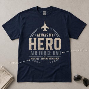 May include: A navy blue short-sleeve t-shirt featuring a graphic print with a central airplane icon, the text ALWAYS MY HERO, AIR FORCE DAD, MICHAEL, and SERVING WITH HONOR, along with two arrows pointing to the text with labels CUSTOM RELATION and CUSTOM NAME.
