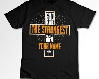 May include: Black t-shirt with an orange cross graphic. The cross has the text "God Made The Strongest Named Them Your Name" on it.