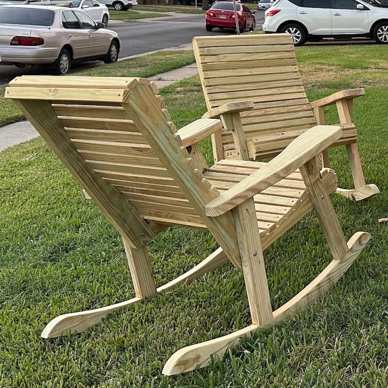 Op de afbeelding: Twee houten schommelstoelen van lichtgekleurd hout staan op een grasveld. De stoelen hebben latten rugleuningen en zittingen, met armleuningen. De schommels zijn gebogen, waardoor een zachte schommelbeweging mogelijk is. De stoelen staan buiten.