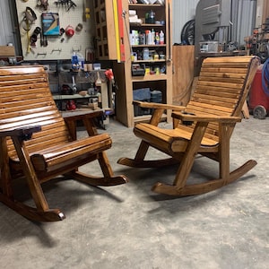 Op de afbeelding: Twee houten schommelstoelen met een glanzende afwerking. De stoelen hebben een lattenrugleuning en zitting, armleuningen en een ingebouwde bekerhouder. De schommelstoelen zijn ontworpen voor comfort en ontspanning.