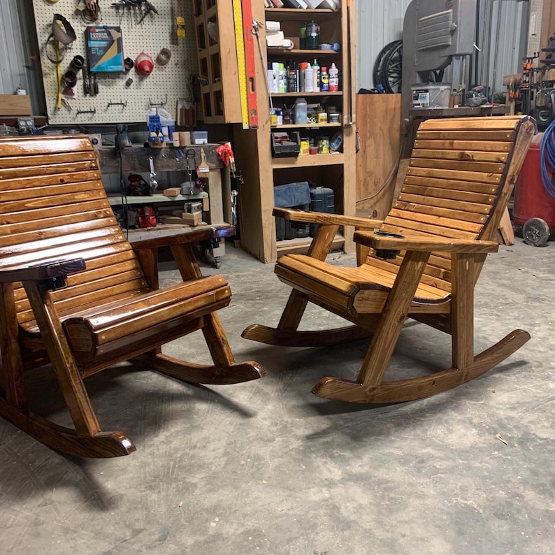 Op de afbeelding: Twee houten schommelstoelen met een glanzende afwerking. De stoelen hebben een lattenrugleuning en zitting, armleuningen en een ingebouwde bekerhouder. De schommelstoelen zijn ontworpen voor comfort en ontspanning.