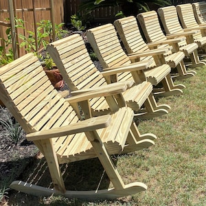 Op de afbeelding: Een rij houten schommelstoelen met lattenrugleuningen en armleuningen. De stoelen zijn gemaakt van lichtgekleurd hout en staan op een gazon, wat duidt op gebruik buitenshuis. Het ontwerp is eenvoudig en klassiek.