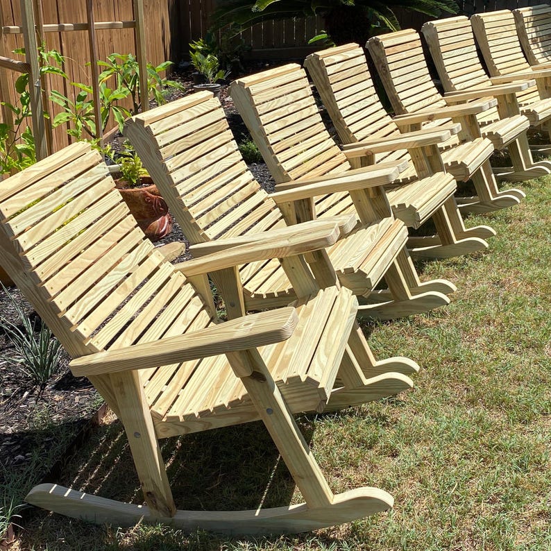 Op de afbeelding: Een rij houten schommelstoelen met lattenrugleuningen en armleuningen. De stoelen zijn gemaakt van lichtgekleurd hout en staan op een gazon, wat duidt op gebruik buitenshuis. Het ontwerp is eenvoudig en klassiek.
