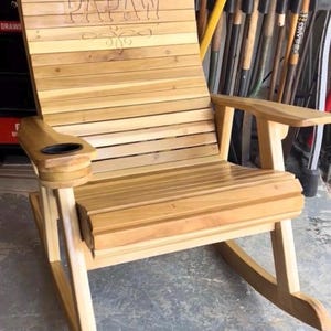 Op de afbeelding: Houten schommelstoel met horizontale lamellen en armleuningen. De rugleuning is voorzien van het gegraveerde woord "PAPAW" met decoratieve krullen. Een bekerhouder is ingebouwd in de armleuning. De stoel is gemaakt van lichtgekleurd hout.