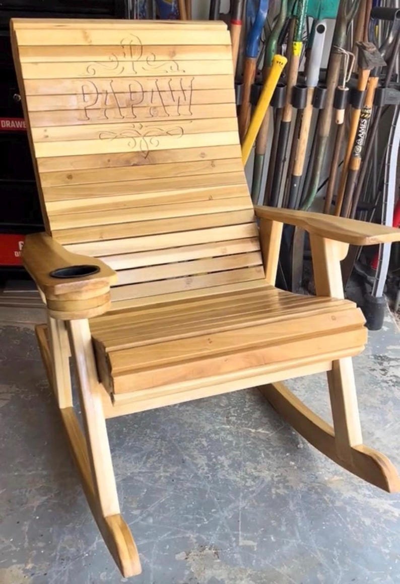 Op de afbeelding: Houten schommelstoel met horizontale lamellen en armleuningen. De rugleuning is voorzien van het gegraveerde woord "PAPAW" met decoratieve krullen. Een bekerhouder is ingebouwd in de armleuning. De stoel is gemaakt van lichtgekleurd hout.