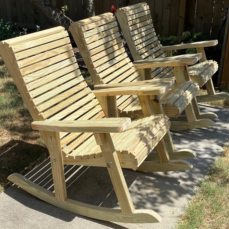 Op de afbeelding: Drie houten schommelstoelen van lichtgekleurd hout. Elke stoel heeft een lattenrugleuning en zitting, armleuningen en gebogen schommelstoelen. De stoelen staan in een rij op een betonnen ondergrond, met een natuurlijke achtergrond.