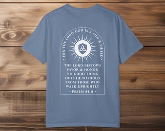 May include: A light blue t-shirt with a white graphic design featuring a sun with a triquetra symbol inside. The text reads "For the Lord God is a Sun & Shield. The Lord bestows favor & honor. No good thing does He withhold from those who walk uprightly. Psalm 84:11."