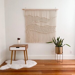 May include: A macrame wall hanging with a woven pattern of waves and a fringe bottom. The hanging is displayed on a white wall with a wooden floor, a small side table, and a potted plant.