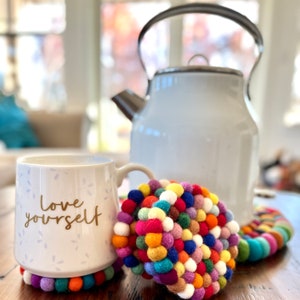 May include: A white ceramic mug with the words "Love yourself" in gold lettering, sitting on a colorful felt coaster. The mug is next to a white enamel teapot.